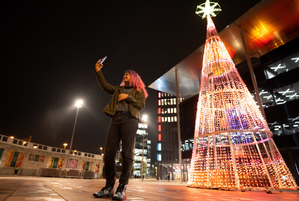 youg woman with pink hair taking a selfie in front of a 12 metre high illuminated pixel christmas tree in central square cardiff