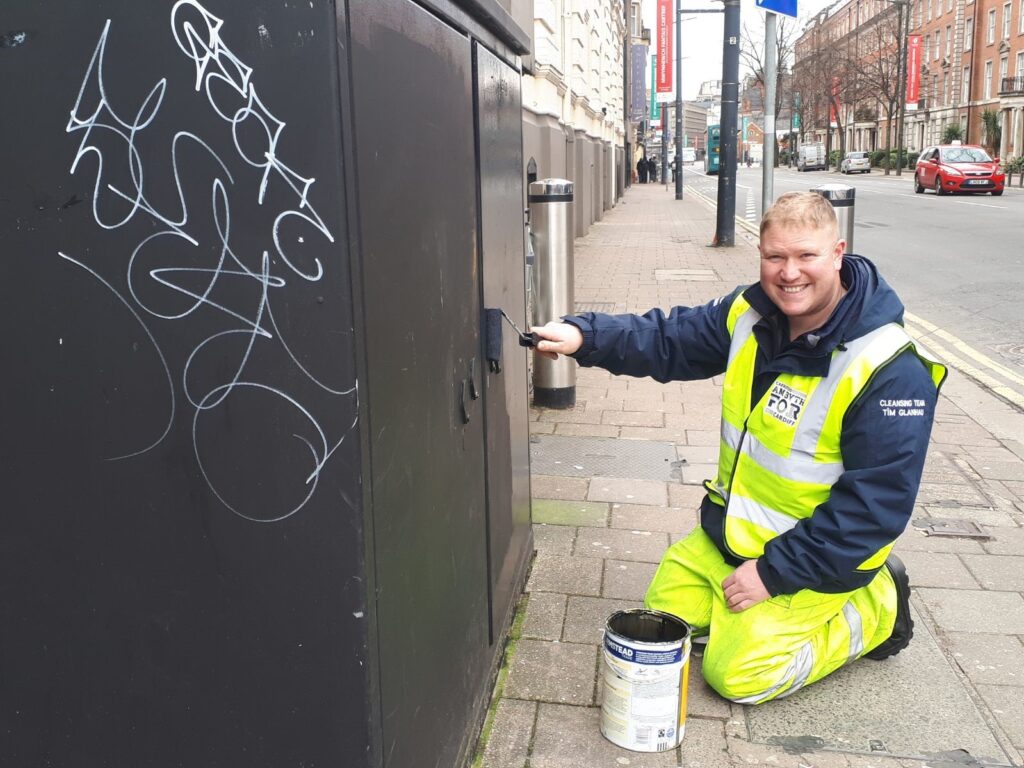man wearing for cardiff cleansing team uniform smiling and painting an electric box