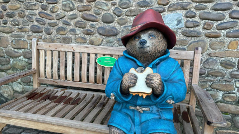 A Paddington bear statue sat on a bench in front of a cobblestone wall eating a marmalade sandwich